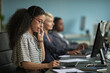 © DragonImages - Young adult woman wearing headset talking on phone and taking notes while working at computer in modern office with two female colleagues in background