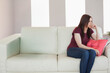 © WavebreakMediaMicro - Woman sitting on cream sofa in living room, holding coral pillow, gazing at windows, copy space