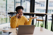© MINAE - Young black female freelancer is talking on phone and gesturing while working on laptop in a cafe, enjoying a cup of coffee and using a tablet