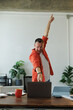 © Tj - Happy businessman pointing at his laptop and raising one arm in the air while celebrating success in his modern home office, expressing excitement and achievement