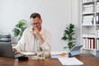 © Tj - Stressed businessman with hand on his chin looking worried at his laptop while calculating home finances sitting at his desk with cash, coins, documents and a tablet
