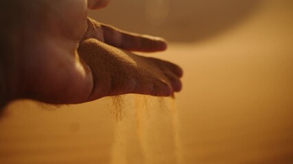 Close-up of golden desert sand slipping gracefully through a hand at sunset in the Sahara Desert, Morocco, with warm light creating a soft glow.