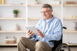 © Prostock-studio - Joyful impaired senior man in wheelchair using tablet computer at home. Positive retired male with physical disability browsing web on touch pad, watching video online indoors