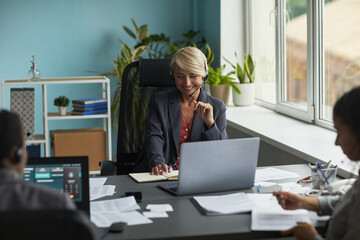 Wall Mural - Caucasian middle aged woman wearing headset smiling while working at desk with laptop and documents, with colleagues working in blurred foreground