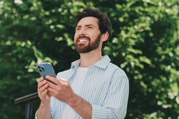 Young man outdoors smiling while using a smartphone, displaying a casual and cheerful look amidst lush greenery.