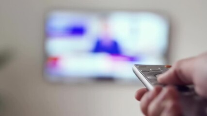 Out-of-focus hand holding a TV remote, with a blurred television screen in the background