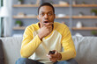 © Prostock-studio - Shocked african american young man in casual outfit with TV remote sitting on couch at home, watching news or horror movie, touching his chin, copy space, living room interior
