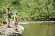 © pressmaster - Middle aged man and boy fishing together on rocky riverbank, both holding fishing rods and focusing on water, lush green trees and flowing river in background
