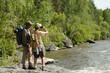 © pressmaster - Middle aged Black man and teenage boy standing on rocky riverbank fishing together, both wearing backpacks, boy holding fishing rod and looking toward water, surrounded by green forest