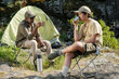 © pressmaster - Middle aged man and teenage boy sitting on folding chairs near tent drinking from metal cups outdoors, engaging in conversation during fishing trip in rocky nature setting