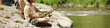 © pressmaster - Middle aged man sitting beside boy fishing with rod on rocky riverbank, man gently placing hand on boy shoulder, both focused on water, outdoor activity