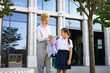 © sofiko14 - A mother helps her daughter with her backpack in front of a school building on a sunny day.