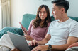 © Daniel - Young Asian couple sitting on green sofa at home, smiling while using laptop and smartphone together. Woman wears pink top, man in white t-shirt with smartwatch, cozy indoor background