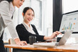 © kenchiro168 - Two Asian businesswomen in formal suits discussing data analysis at modern office desk, smiling and collaborating on computer with business growth chart, teamwork and productivity