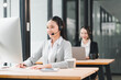 © kenchiro168 - Asian woman customer service representative wearing headset and business suit working at computer in modern office, smiling and providing support, with colleague in background