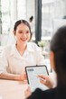 © kenchiro168 - Young woman smiling during job interview with recruiter holding resume on digital tablet in modern office
