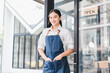 © kenchiro168 - Young Asian woman wearing denim apron stands smiling confidently at entrance of modern cafe, welcoming customers with friendly and inviting expression