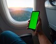 © ZaynR - Man sitting by an airplane window holding a smartphone with a green screen, perfect for mockups and travel concepts during a scenic flight