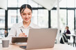 © kenchiro168 - Young Asian woman smiling while working on laptop in modern office, holding pen and clipboard, natural light, professional and focused atmosphere