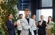 © Studio Romantic - Portrait of a young happy smiling confident bearded business man in formal clothes looking cheerful at camera with team of company employees applauding on background in modern office.