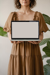 Wall Mural - Woman in Earthy-Toned Dress Presenting Laptop with Blank Screen, Cozy and Natural Setting