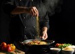 © Roushon - A professional chef in a black uniform is cooking spaghetti with tomato and vegetables in a pan, sprinkling seasoning on the dish in a dark studio kitchen, showcasing culinary skill and motion