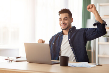 Emotional arab guy freelancer celebrating successful business deal, sitting at workdesk with laptop and raising fist up, copy space, office interior. Young arab man working from home