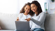 © Prostock-studio - Excited african american mother and kid using laptop together while sitting on couch at home, closeup. Loving black mom and daughter playing online games on computer and hugging, copy space
