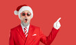 © Studio Romantic - Look here. Funny man in Christmas costume pointing aside. Happy surprised man in suit, tie, Santa cap, white gloves and sunglasses standing on red studio background and pointing index finger to side