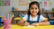 © SimpleDesignStudio - Happy young Indian girl student sitting at desk writing in notebook in classroom. Cute elementary school child learning, doing homework and smiling at camera.