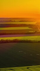 Wall Mural - Bright orange sun setting over endless green crop fields, bathing the rural countryside landscape in a warm golden hour glow -vertical aerial time lapse