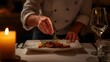 © Ajigus - A chef's hands delicately arrange a gourmet dish, beautifully presented on a fine china plate, highlighting culinary artistry in a warm, intimate dining setting.
