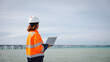 © Happy Photo - A safety-conscious engineer stands at the edge of the water, checking project details on a tablet. The cloudy sky reflects a calm day at the coastal site, emphasizing professionalism