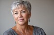 © SerPak - A close-up portrait of a mature woman with short gray hair, wearing earrings and a sweater, exuding confidence and warmth in a neutral background.