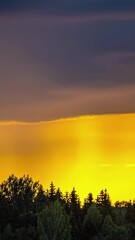 Wall Mural - Timelapse of dramatic golden sunset sky with dark clouds moving fast hiding rays of orange light over forest, vertical