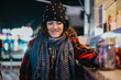 © qunica.com - A young woman wearing winter attire smiles warmly while standing outdoors during a festive holiday evening, surrounded by vibrant lights and good spirits.