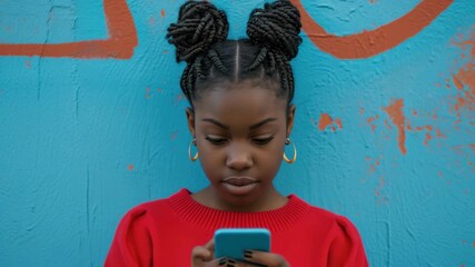 Wall Mural - A young woman with pigtails engaged in texting on her cellphone. She is leaning against a colorful wall which suggests an urban, possibly vibrant and creative community.