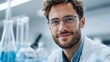 © Yevheniiya - A handsome scientist in a lab coat poses in a laboratory setting, smiling at the camera with a friendly expression. Blue liquid is in the test tubes.