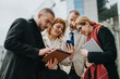 © qunica.com - Businesspeople interacting with technology devices during a meeting. Collaboration, teamwork, and communication emphasized in an outdoor urban setting, demonstrating partnership and brainstorming.