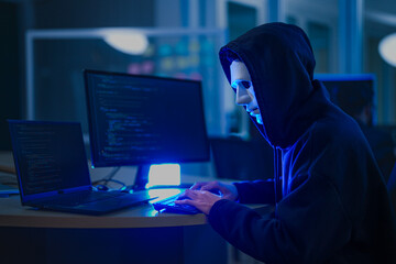 A mysterious hooded hacker working on a computer in a dark room, illuminated by blue screen light. A concept for cybersecurity, hacking, cybercrime, and data privacy threats.