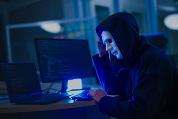 A mysterious hooded hacker working on a computer in a dark room, illuminated by blue screen light. A concept for cybersecurity, hacking, cybercrime, and data privacy threats.