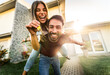 © Davide Angelini - Happy young couple holding home keys after buying real estate - Husband and wife standing outside in front of their new house