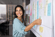 © fizkes - Smiling Latina professional woman placing sticky notes on large planning board, engaged in strategic planning, creative brainstorm session or workflow organization. Project management in modern office