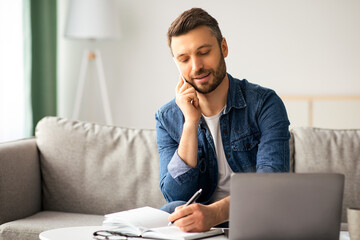 Handsome man freelancer working from home, using laptop and having conversation with clients on mobile phone. Smiling bearded man sitting on couch in living room, talking on smartphone, copy space