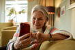 © Marko Geber - Smiling senior woman using smartphone while relaxing at home
