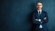 © ittipol - A confident businessman stands with arms crossed, showcasing a professional demeanor. He is wearing glasses and a tailored suit, set against a dark, textured background.