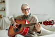 © Seventyfour - Senior Caucasian woman smiling while playing acoustic guitar, sitting on sofa in bright living room, wearing glasses and scarf, enjoying music, showing relaxed and joyful expression