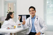 © amnaj - Smiling doctor showing thumbs up gesture of approval while his colleague is working on a computer in the background