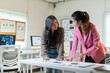© amnaj - Two asian businesswomen leaning over a desk, examining financial reports and discussing company strategy in a modern office