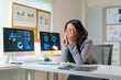 © amnaj - Asian businesswoman covering her face with her hands, suffering from headache or burnout, sitting at desk with financial charts on computer screen